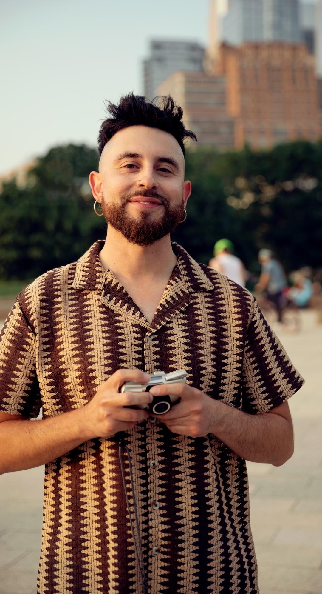 Bearded man in patterned shirt holding a camera, standing outdoors with trees and buildings in the background.