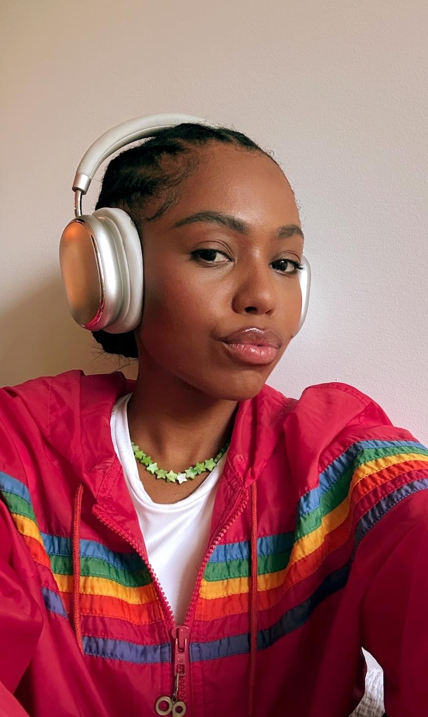 Woman in red rainbow-striped jacket and silver headphones, wearing a green star necklace, looking at the camera.