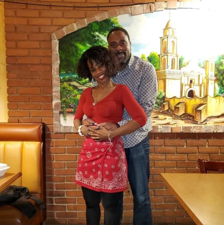 Nyla and Jeff stood smiling at the camera. Nyla is stood in front of Jeff and Jeff has his hands around Nyla's waist and Nyla is holding them.