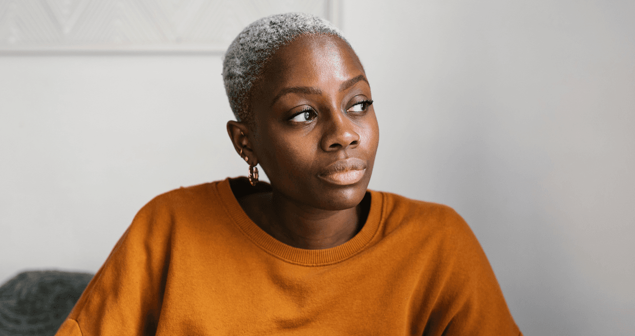 Woman looks out of window thoughtfully. She is wearing an orange jumper.