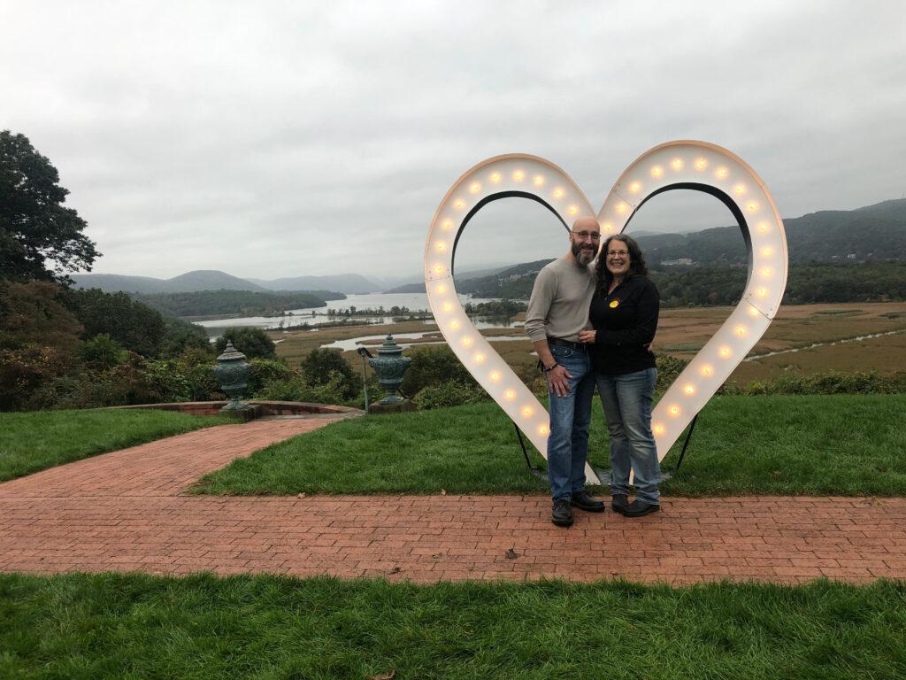 Scott and Wendy stood in front of a large, lit-up, heart-shaped structure in front of a scenic background.