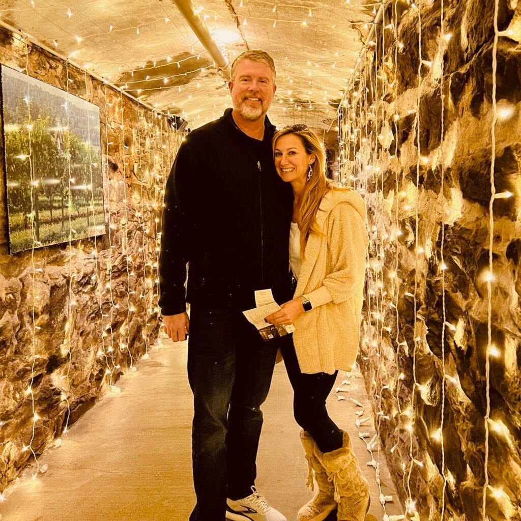 Jim and Meredith standing in a hallway covered in fairy lights and smiling at the camera.