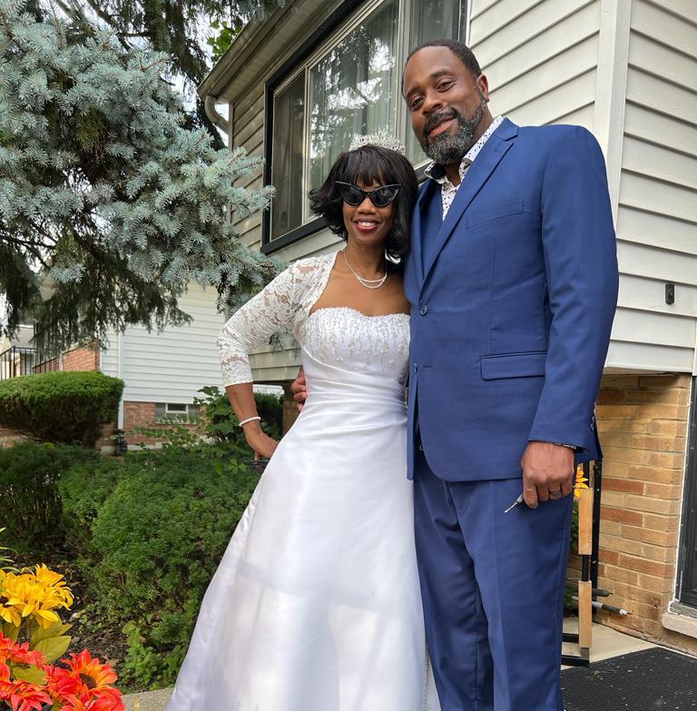 Nyla and Jeff stood with their arms around each other and smiling at the camera on their wedding day. Nyla is wearing a white wedding dress and Jeff is wearing a dark blue suit. 